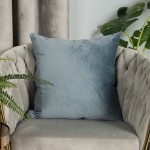 Poszewka welwetowa 40x40 w kolorze błękitnym, umieszczona na szaro-beżowym, tuftowanym fotelu. Wykonana z miękkiego materiału, z delikatną fakturą. Otoczona zielonymi liśćmi roślin, co tworzy przytulny klimat.