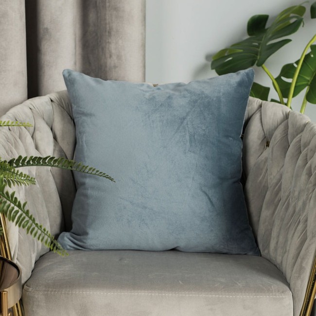Poszewka welwetowa 40x40 w kolorze błękitnym, umieszczona na szaro-beżowym, tuftowanym fotelu. Wykonana z miękkiego materiału, z delikatną fakturą. Otoczona zielonymi liśćmi roślin, co tworzy przytulny klimat.