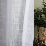 Firana na przelotkach w kolorze białym, wykonana z etaminy, gładka, o wymiarach 140x250 cm. Widoczna faktura materiału oraz delikatny wzór. Zawieszona obok doniczki z rośliną, w aranżacji wnętrza.