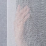 Firana na przelotkach, gładka, w kolorze białym, wykonana z etaminy. Ujęcie z bliska, z widoczną ręką dotykającą materiału, podkreślającą jego lekkość i transparentność. Wymiary 140x250 cm.