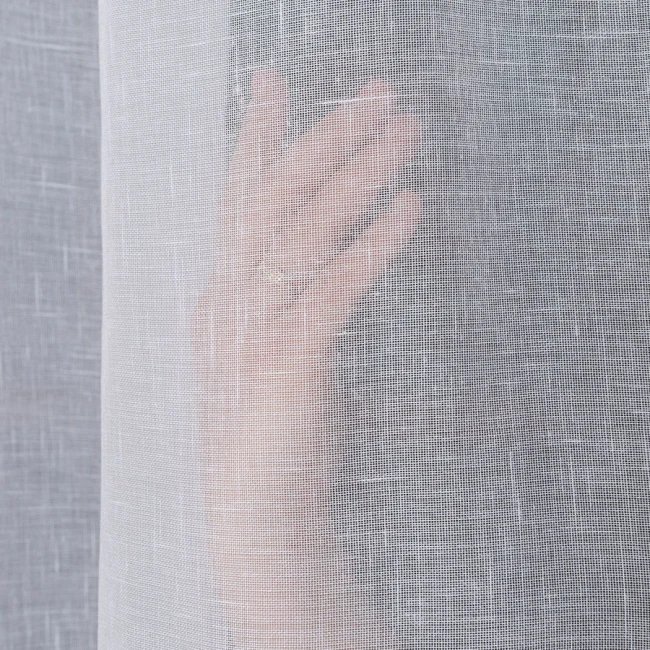 Firana na przelotkach, gładka, w kolorze białym, wykonana z etaminy. Ujęcie z bliska, z widoczną ręką dotykającą materiału, podkreślającą jego lekkość i transparentność. Wymiary 140x250 cm.