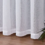 Firana na przelotkach w kolorze białym, wykonana z etaminy, o wymiarach 140x250 cm. Zbliżenie na dolną część firany z widocznym wykończeniem, na tle drewnianej podłogi oraz rośliny w prawym dolnym rogu.