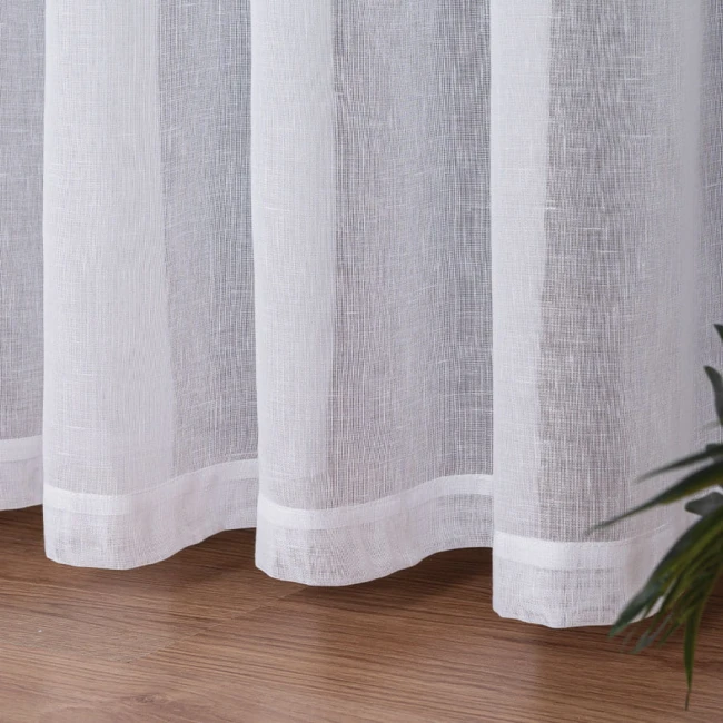 Firana na przelotkach w kolorze białym, wykonana z etaminy, o wymiarach 140x250 cm. Zbliżenie na dolną część firany z widocznym wykończeniem, na tle drewnianej podłogi oraz rośliny w prawym dolnym rogu.