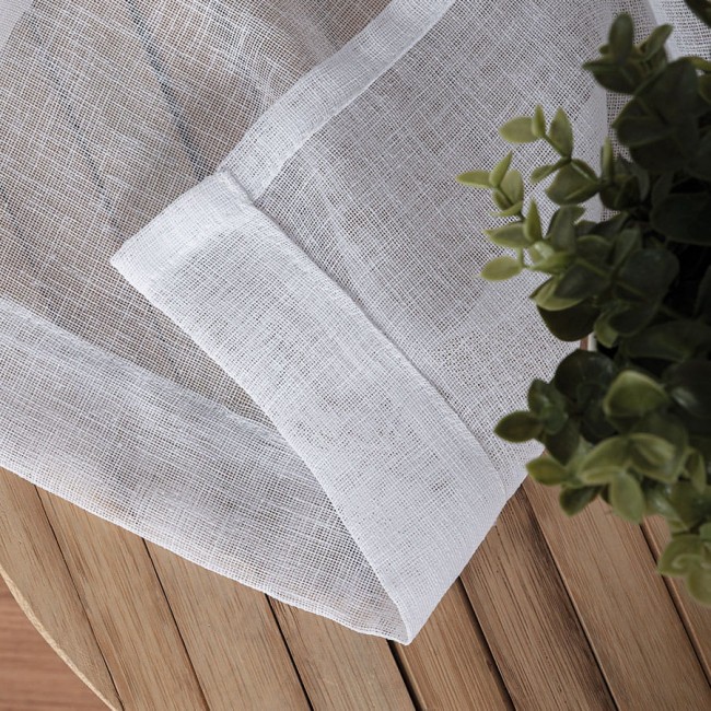 Biała firana na przelotkach z etaminy, gładka, o wymiarach 140x250 cm. Ujęcie z bliska, pokazujące teksturę materiału oraz wykończenie brzegów. W tle widoczna roślina doniczkowa, na jasnej drewnianej powierzchni.
