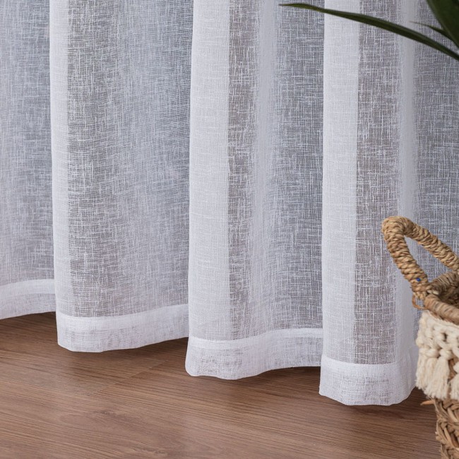 Firana na przelotkach gładka, biała z etaminy, o wymiarach 140x250 cm. Widoczna tekstura materiału, delikatnie przepuszczającego światło. Ujęcie z boku, z ułożonym dołem. W tle widać pleciony kosz.