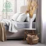 Prześcieradło frotte z gumką 90x200 w kolorze popielatym, rozłożone na łóżku z jasnymi poduszkami. Kąt ujęcia frontalny, z elementami aranżacji wnętrza, jak stół nocny z białym wazonem i dekoracją z traw zielonych na tle jasnych zasłon.