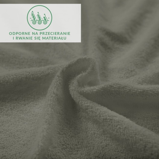 Prześcieradło frotte 160x200 w kolorze szarym, rozłożone na podłożu, z widoczną teksturą materiału. Zbliżenie na miękką, elastyczną tkaninę, odporną na przetarcia oraz rwanie. W prawym górnym rogu zielona ikona informacyjna.