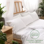 Prześcieradło jersey z gumką w kolorze białym o wymiarach 140x200, leżące na łóżku z poduszkami. Widoczna gładka faktura materiału oraz jasne otoczenie. W tle drewniane łóżko i roślina doniczkowa. Logo z napisem IDEALNIE PRZYLEGA DO MATERACA.