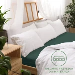 Prześcieradło jersey z gumką w kolorze zieleni butelkowej, w rozmiarze 90x200 cm, idealnie przylega do materaca. Widoczne białe poduszki i delikatna pościel na tle drewnianego łóżka oraz roślinność w otoczeniu.