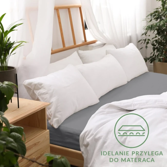Prześcieradło jersey z gumką w kolorze szarym o wymiarach 200x220 cm, idealnie dopasowane do materaca. Ułożone na łóżku z białymi poduszkami, w otoczeniu rośliny. Tło jasne i przytulne, podkreślające teksturę materiału.