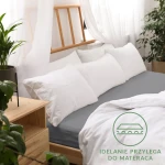 Prześcieradło jersey z gumką 140x200 w kolorze szarym leży na łóżku, otoczone białymi poduszkami. Widoczne logo z napisem Idealnie przylega do materaca. Tło jasne, z roślinnością i drewnianymi meblami.