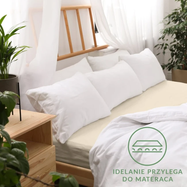 Prześcieradło jersey z gumką w kolorze jasnego beżu, rozmiar 200x220 cm, leży na łóżku z białą pościelą i poduszkami. Widoczny detal gumki, która zapewnia idealne przyleganie do materaca. Otoczenie w jasnym, przytulnym wnętrzu.