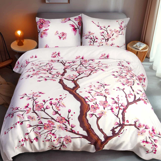 Pościel satynowa 180x200 w kolorze białym z motywem drzewa wiśniowego. Zestaw zawiera poszewki na poduszki z tym samym wzorem. Ułożona na łóżku, w tle widoczna nocna lampka i stolik.