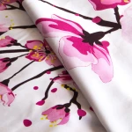 Pościel satyna bawełniana 180x200 w białym kolorze z nadrukiem różowych kwiatów wiśni. Widoczne detale kwiatów oraz gałęzi na tle. Materiał gładki, z lekkim połyskiem, ułożony w lekkim zagięciu.