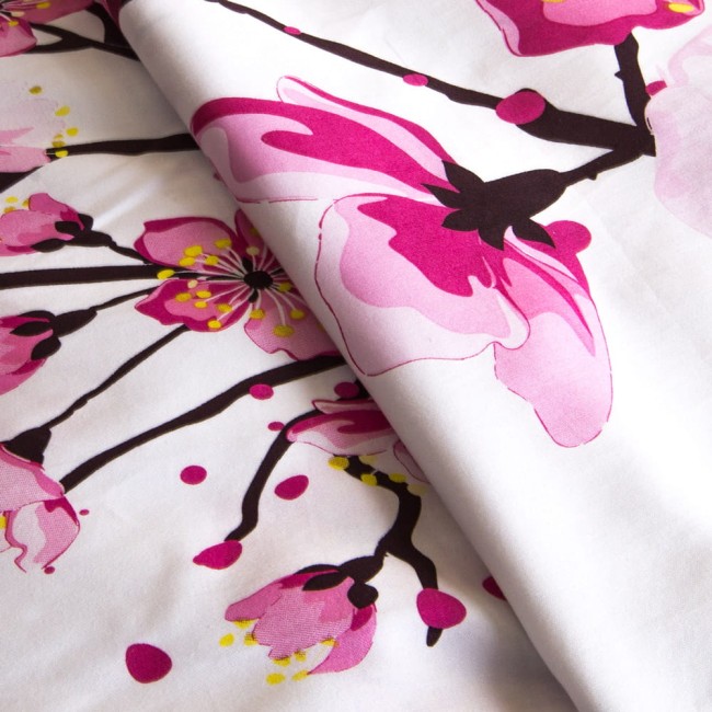 Pościel satyna bawełniana 180x200 w białym kolorze z nadrukiem różowych kwiatów wiśni. Widoczne detale kwiatów oraz gałęzi na tle. Materiał gładki, z lekkim połyskiem, ułożony w lekkim zagięciu.