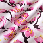 Pościel satyna bawełniana 180x200 z białym tłem ozdobionym różowymi kwiatami wiśni i ciemnymi gałęziami. Widoczne detale kwiatów z żółtymi pręcikami, materiał gładki, w zbliżeniu na wzór.