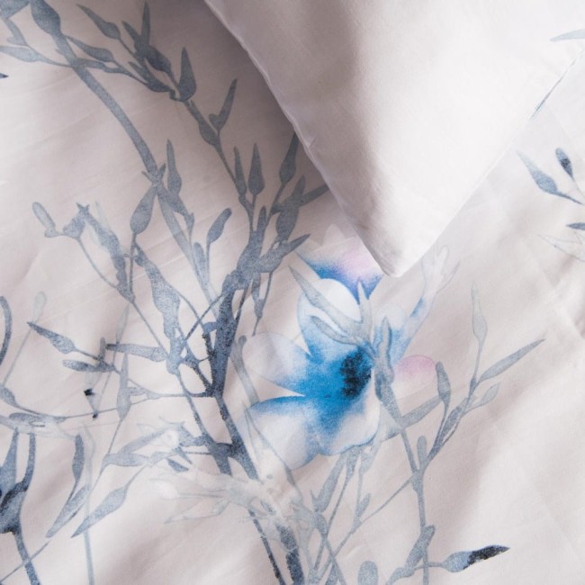 Pościel satyna bawełniana 220x200 w kolorze jasnym, z pastelowymi, błękitnymi kwiatkami. Złożona, z widocznym nadrukiem roślinnym na materiale. W górnej części widoczna poszewka na poduszkę. Tło białe, co podkreśla delikatność wzoru.