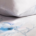 Pościel satynowa 220x200 cm w jasnym kolorze z pastelowymi błękitnymi kwiatkami. Zbliżenie na poduszkę leżącą na poszewce, ukazujące subtelny wzór oraz gładką fakturę materiału. Tło jednolite, co podkreśla detale produktu.