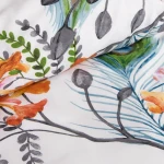 Pościel bawełniana 140x200 Cottonlove w kolorze białym, zdobiona tropikalnymi pomarańczowymi kwiatami oraz zielonymi liśćmi. Pozycjonowana w rozłożonej formie, zbliżenie na detale wzoru, na jasnym tle. Tekstura tkaniny gładka.