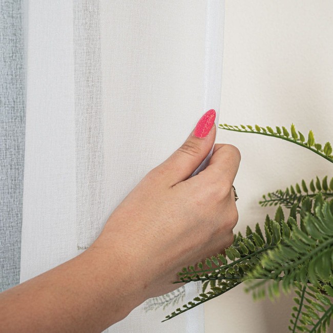 Firana gotowa na srebrnych przelotkach, biała, z faktury półprzezroczystej tkaniny, wisząca na ścianie. Ręka z różowym lakierem do paznokci delikatnie trzyma firanę. W tle widoczna zieleń rośliny doniczkowej, na jasnym tle. Wymiary: 140x250 cm.