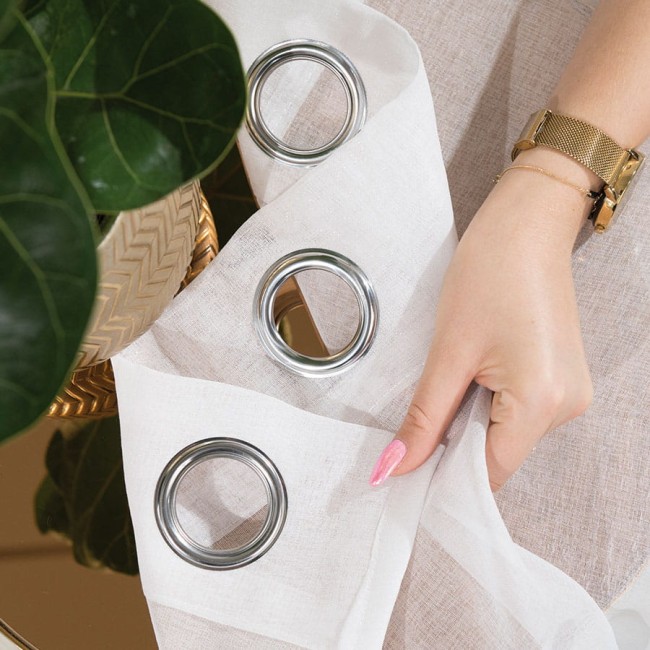 Firana w kolorze białym na srebrnych przelotkach, o wymiarach 140x250 cm. Widoczna w ręce, delikatna faktura materiału. Srebrne kółka na górnej krawędzi. Tło w jasnych odcieniach z rośliną w doniczce.