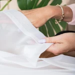 Firana gotowa w kolorze białym, wykonana z lekkiego materiału woalu, z widocznymi srebrnymi przelotkami. Ujęcie z bliska, ukazujące detale materiału i wykończenia, z wyraźnie zarysowanymi brzegami. Tło neutralne, zielona roślina w tle.