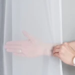 Firana gotowa na srebrnych przelotkach, w kolorze białym, o wymiarach 140x250 cm, wisząca, wykonana z lekkiego materiału woalowego. Zdjęcie przedstawia rękę odsłaniającą firanę, ukazującą jej przezroczystość i delikatną teksturę. Tło szare.