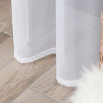 Firana gotowa w białym woalu na srebrnych przelotkach o wymiarach 140x250 cm, zawieszona, z delikatną teksturą, lekko przepuszczająca światło. Widoczny dolny kant firany z estetycznym wykończeniem. Tło drewniana podłoga.