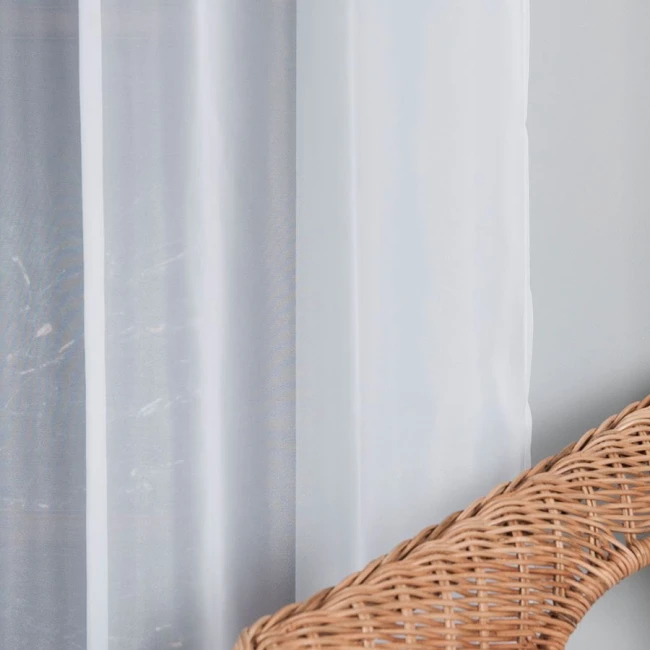 Firana woalowa w kolorze białym, o wymiarach 140x250 cm, zawieszona na srebrnych przelotkach. Produkt widoczny z boku, w tle ściana, na pierwszym planie pleciona rattanowa oprawa.