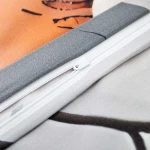 Zbliżenie na zamek błyskawiczny pościeli satynowej w szarym kolorze z pomarańczowymi kwiatami, pokrytej gładkim materiałem. Widoczne fragmenty wzoru oraz kolorowe detale na tkaninie, material pokryty subtelną teksturą.