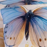 Poszewka na poduszkę z motywem niebieskiego motyla na kremowym tle. Wzór przedstawia detale skrzydeł z gradientem kolorów, widoczny zbliżenie.