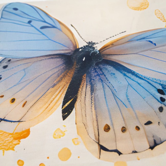 Kremowa pościel satyna bawełniana 180x200 z niebieskimi motylami, zbliżenie na motyla. Widoczne błękitno-niebieskie skrzydła i czarne detale, wzór na tkaninie z delikatnymi akcentami w postaci kropek. Tło gładkie, jasne.