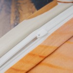 Zbliżenie na zamek błyskawiczny pościeli satynowej w kremowym kolorze, z widoczną fakturą materiału w odcieniach beżu i brązu. Zamek umiejscowiony na krawędzi poszewki, wskazuje na sposób zamykania.