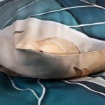 Poduszka z pościeli satynowej w kolorach turkusowym i złotym, z delikatnym wzorem liści. Ujęcie z bliska podkreśla fakturę tkaniny i okrągły kształt poduszki. Widoczne są także płynne przejścia kolorystyczne. W tle widoczny fragment poszwa.
