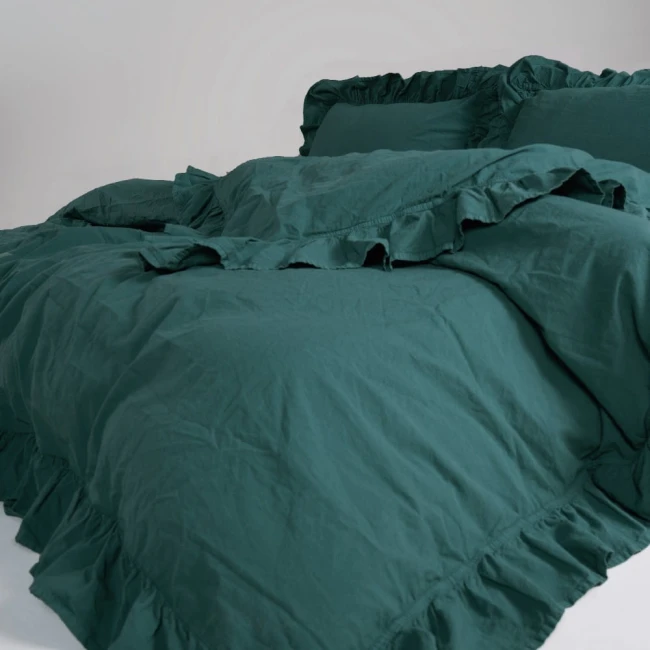 Pościel bawełniana z falbanką w kolorze butelkowej zieleni, rozmiar 220x200 cm, leży na łóżku z widoczną falbanką na krawędzi narzuty i poduszek. Tło jest jasne i jednolite, podkreślające teksturę materiału. Zestaw składa się z narzuty i dwóch poduszek.