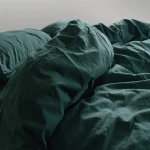 Pościel bawełniana w kolorze butelkowej zieleni, rozłożona na łóżku, z falbanką. Widać poduszki oraz kołdrę o wymiarach 220x200 cm. Tekstura materiału jest lekko pomarszczona, co podkreśla naturalny styl. Tło jasnoszare.