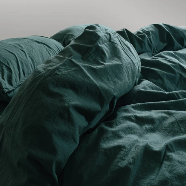 Pościel bawełniana w kolorze butelkowej zieleni, rozłożona na łóżku, z falbanką. Widać poduszki oraz kołdrę o wymiarach 220x200 cm. Tekstura materiału jest lekko pomarszczona, co podkreśla naturalny styl. Tło jasnoszare.