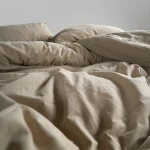 Pościel bawełniana z falbanką 160x200 Stonewashed Exclusive w odcieniu beżowym, leżąca na łóżku. Widoczne są poduszki oraz narzuta z miękkim wykończeniem. Tło jasne, co podkreśla teksturę materiału. Zbliżenie na falbany i naturalne marszczenia.