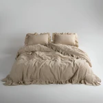 Pościel bawełniana z falbanką 160x200 w odcieniu beżowym, rozłożona na białym tle. Zawiera dwie poszewki z falbanami oraz narzutę z falbanką na brzegach, ukazując delikatną fakturę materiału. Widok frontalny ukazuje całość zestawu.