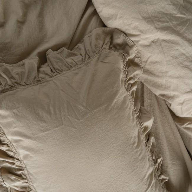 Pościel bawełniana z falbanką w kolorze beżowym, rozmiar 160x200. Zbliżenie na poszewkę z falbanką i prześcieradło, ułożone na łóżku. Tekstura materiału jest miękka i lekko marszczona. Prosta, neutralna aranżacja.