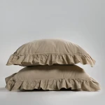 Pościel bawełniana z falbanką w kolorze beżowym, rozmiar 160x200. Na zdjęciu widoczne dwa poduszki, leżące jedna na drugiej. Materiał jest gładki, a falbanka wykończona na dolnej krawędzi poduszek. Tło jednolite, szare.