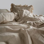 Pościel bawełniana w kolorze beżowym, z falbanką, rozłożona na łóżku. Zawiera poszewki na poduszki i narzutę z delikatnymi krawędziami. Materiał o fakturze stonewashed, odpowiedni do standardowego łóżka 220x200. Tło proste, jasne.