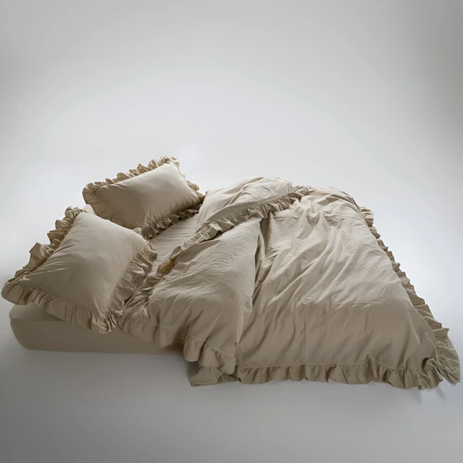 Pościel bawełniana w kolorze beżowym, rozmiar 220x200, z falbanym wykończeniem. Widoczna na łóżku w ułożeniu leżącym, obejmująca poduszki z falbanką. Tło proste, jasne, podkreślające detal materiału.
