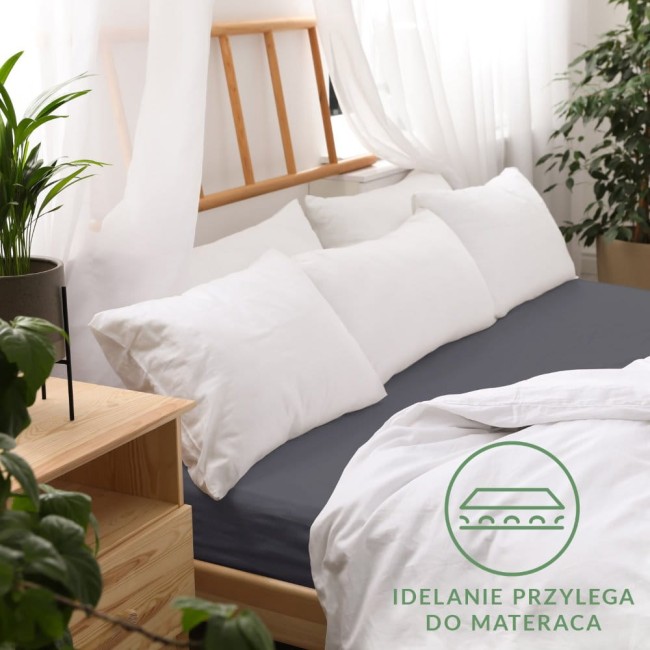 Prześcieradło jersey z gumką 140x200 w kolorze ciemnoszarym, rozłożone na łóżku, idealnie przylegające do materaca. W tle białe poduszki i drewniana rama łóżka. Proste tło z roślinnością oraz wręcz ciepła, przytulna aranżacja sypialni.