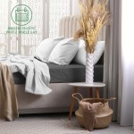 Prześcieradło frotte z gumką 160x200 w kolorze ciemno-szarym, leżące na łóżku z białymi poduszkami. Tło to elegancka sypialnia z jasnymi zasłonami i dekoracyjną ścianą. Widoczny stół nocny oraz kosz z dodatkowymi tekstyliami.