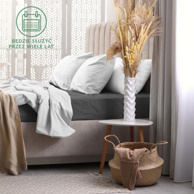 Prześcieradło frotte z gumką 160x200 w kolorze ciemno-szarym, leżące na łóżku z białymi poduszkami. Tło to elegancka sypialnia z jasnymi zasłonami i dekoracyjną ścianą. Widoczny stół nocny oraz kosz z dodatkowymi tekstyliami.