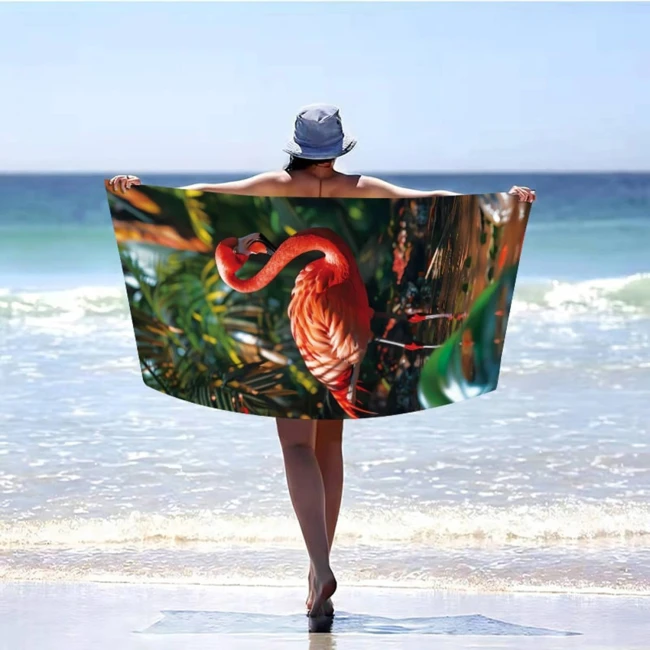 Ręcznik plażowy z mikrofibry o wymiarach 100x180 cm, w różowym kolorze z wzorem flaminga na zielonym tle. Ułożony na plaży, podnoszony przez osobę w kapeluszu. Produkt szybkoschnący, z dużą grafiką, idealny na plażę lub basen.