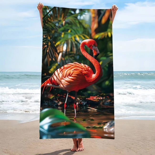 Ręcznik plażowy 100x180 cm z mikrofibry w różowym kolorze, wizualizujący flaminga na tle tropikalnej roślinności. Produkt rozłożony, trzymany w dłoniach, ukazuje szczegóły wzoru flaminga oraz fakturę materiału. Tło z morzem i piaskiem.
