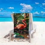 Ręcznik plażowy 100x180 cm w kolorze różowym z mikrofibry. Wzór przedstawia flaminga na tle tropikalnej roślinności. Produkt leży rozwinięty na leżaku, podkreślając swój duży rozmiar oraz szybkoschnącą tkaninę. Tło to plaża z błękitnym morzem.