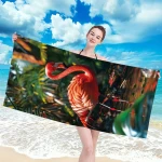 Ręcznik plażowy 100x180 cm z mikrofibry w dużym rozmiarze, różowy z wzorem flaminga. Produkt trzymany przez uśmiechniętą osobę na tle plaży z palmami i morzem, rozłożony. Wysoka jakość tkaniny, szybkoschnący materiał idealny na plażę.